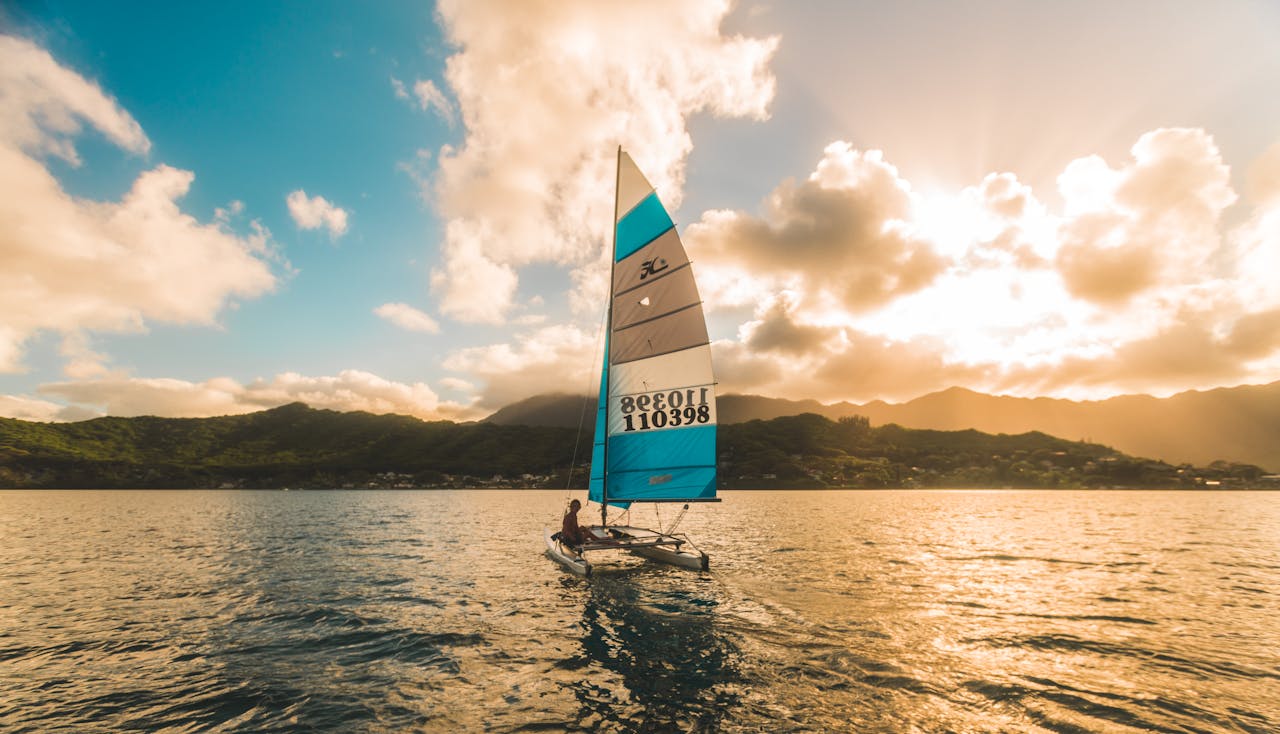 A sailboat glides on Kaneohe Bay at sunset with mountains in the background.