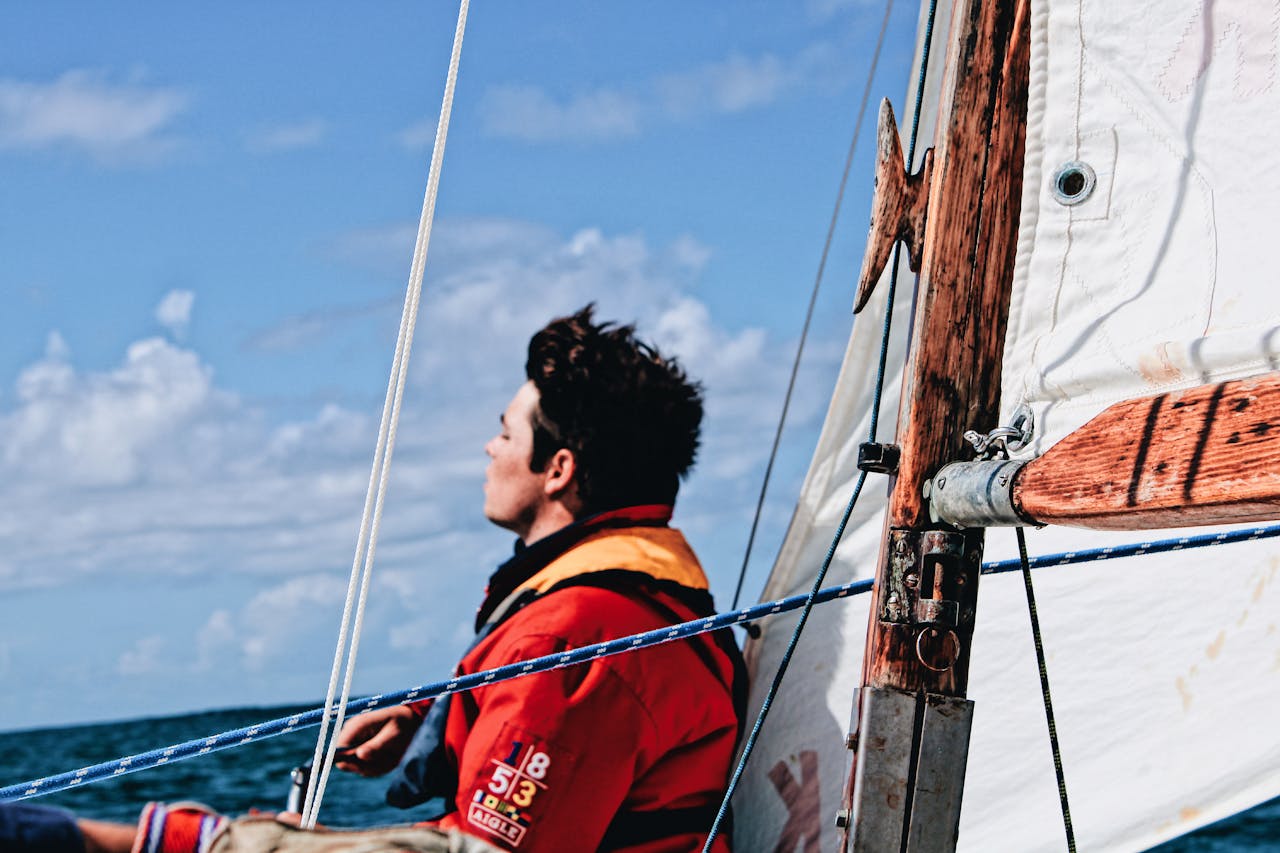 A solitary sailor in a red jacket gazes at the sky while navigating a sailboat under clear blue skies.